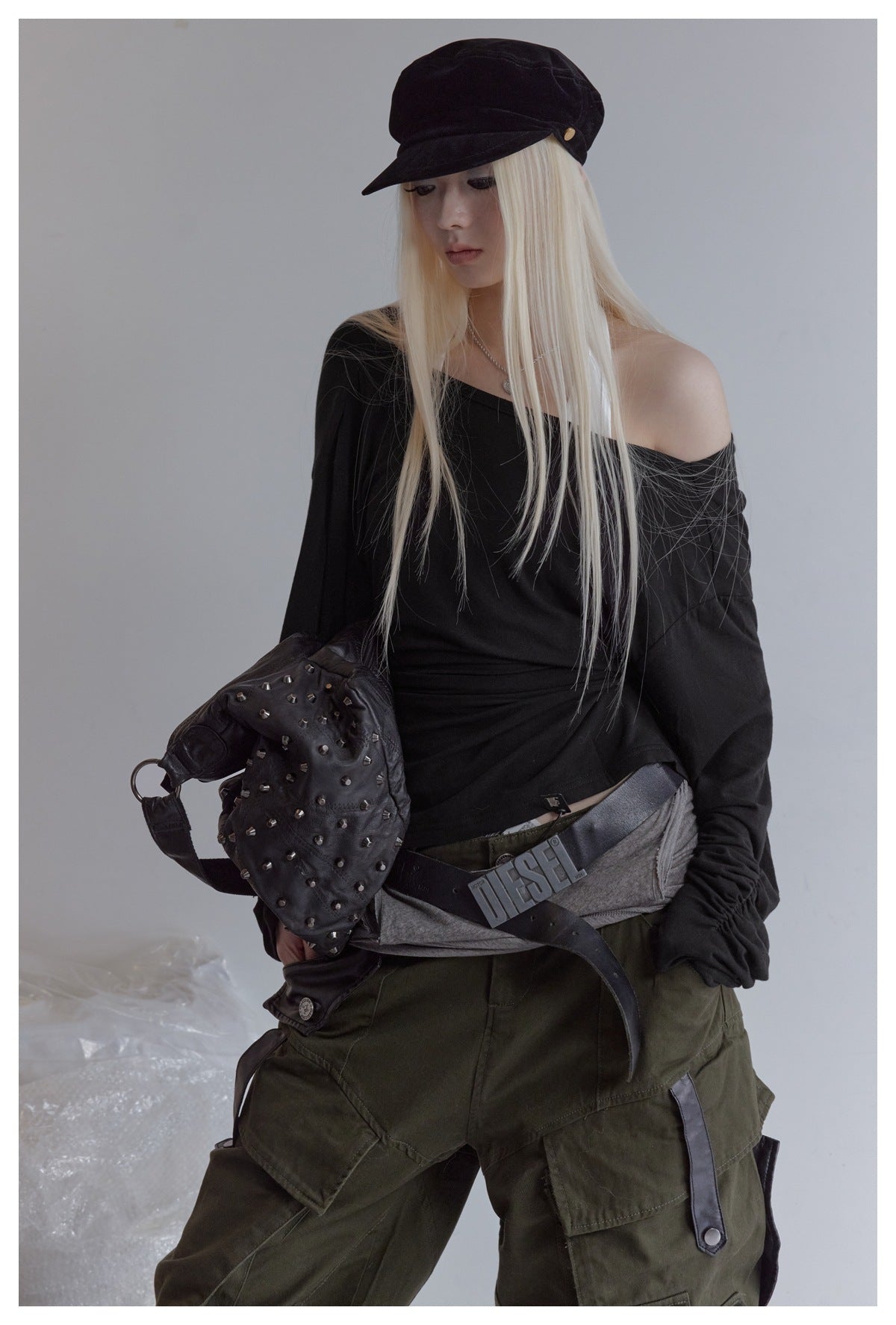 Model wearing the black draped long-sleeve top off one shoulder with a velvet cap, studded bag, belt and olive cargo pants