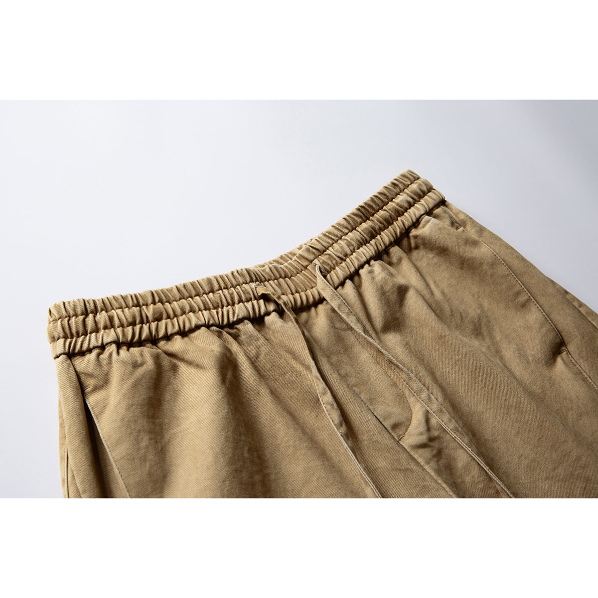 Close-up of tan trousers showing the gathered elastic waistband, drawstring and angled front pocket.