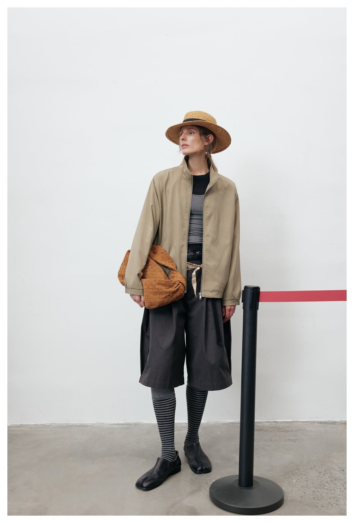 Model wearing an open khaki zip jacket over a gray top with dark shorts, straw hat and woven tan bag
