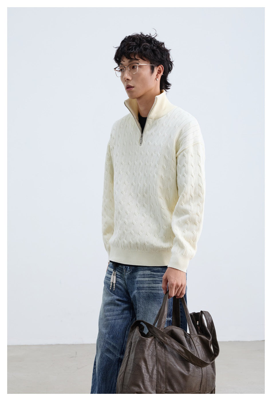 Model wearing an ivory cable-knit quarter-zip sweater with a raised collar and blue jeans, holding a dark tote bag.
