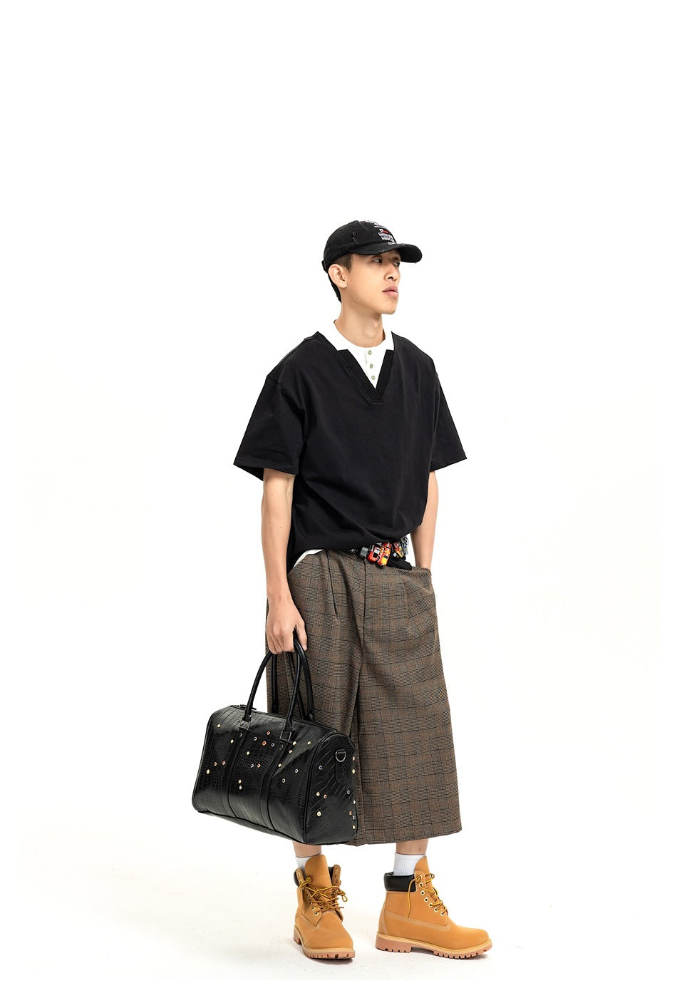 Model wearing a black V-neck short-sleeve top, plaid midi skirt, black cap, and tan boots, carrying a black bag.