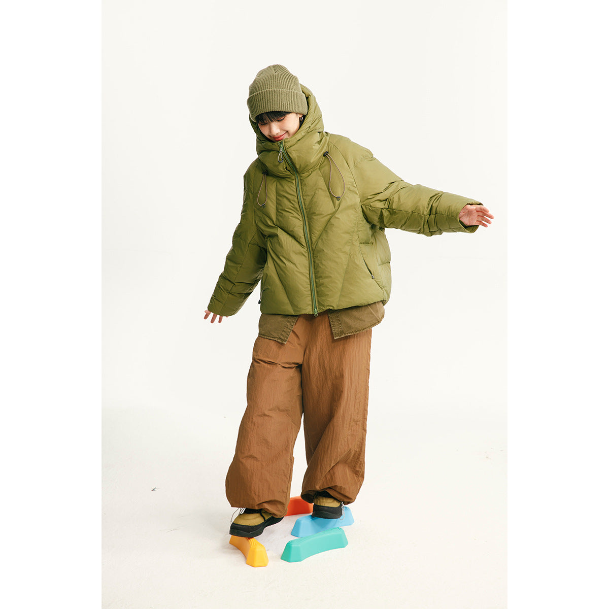 Model wearing an olive hooded puffer jacket and brown wide-leg pants while balancing on colorful blocks.