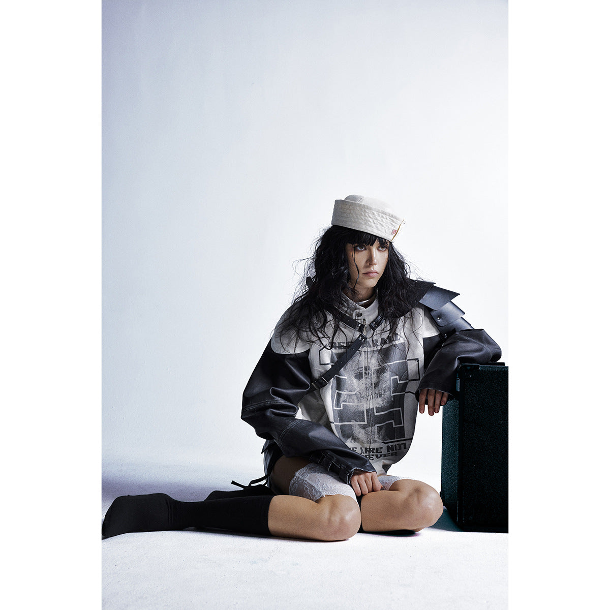 Model seated in a cream-and-black graphic jacket with a white cap, black skirt, lace thigh-highs and black socks.