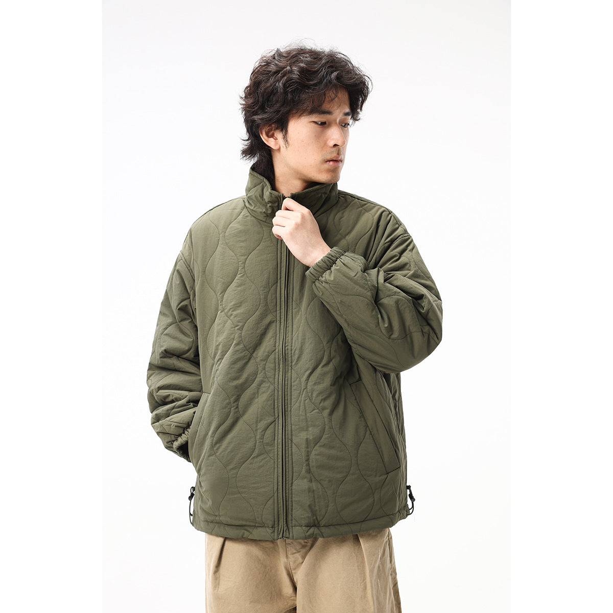 Man wearing an olive quilted jacket and holding its raised collar, showing the front zipper, pockets and stitching.