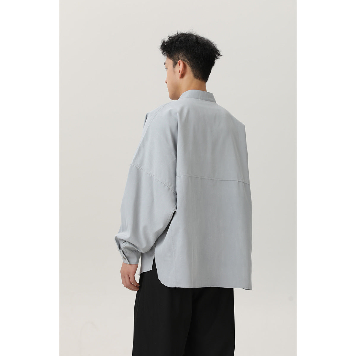 Rear view of a model wearing a light gray oversized shirt with a stand collar, dropped sleeves, and a horizontal back seam.
