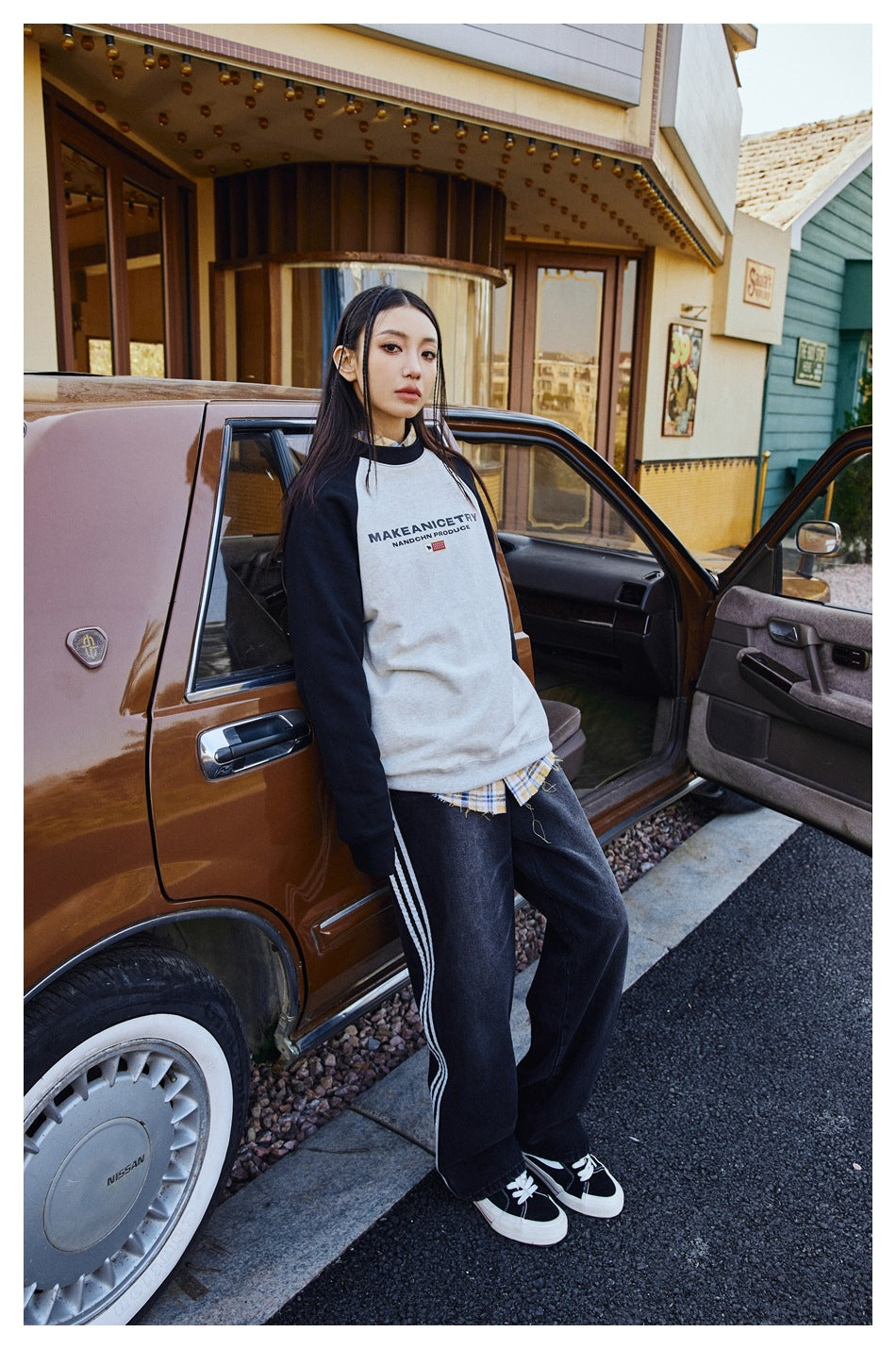 A model wears a cream raglan sweatshirt with black sleeves and dark striped pants beside a car.