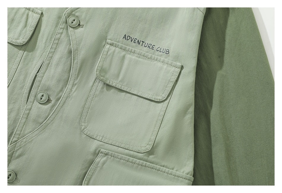 Close-up of a sage-green button-front garment showing a flap chest pocket with “Adventure Club” lettering.