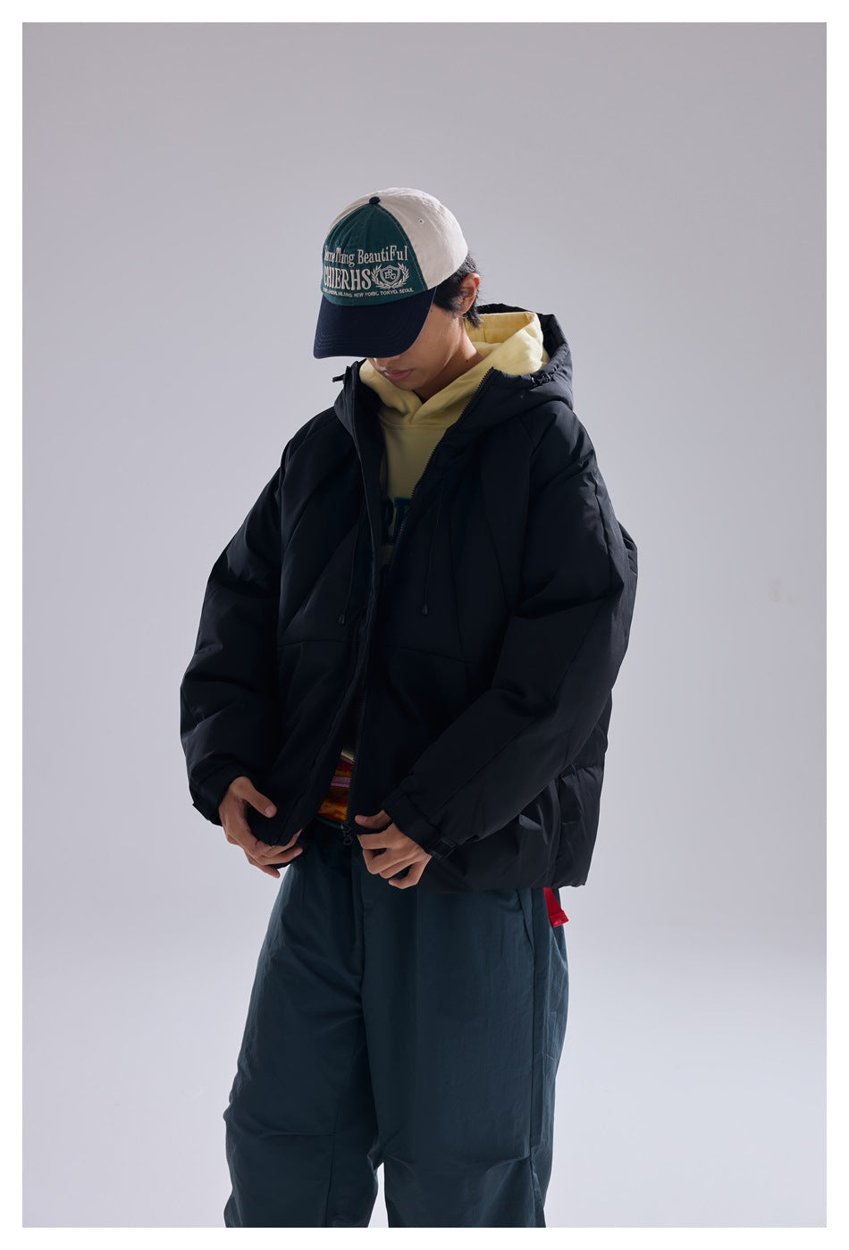 Model wearing a black hooded puffer jacket over a yellow hoodie, looking down and holding the jacket front.
