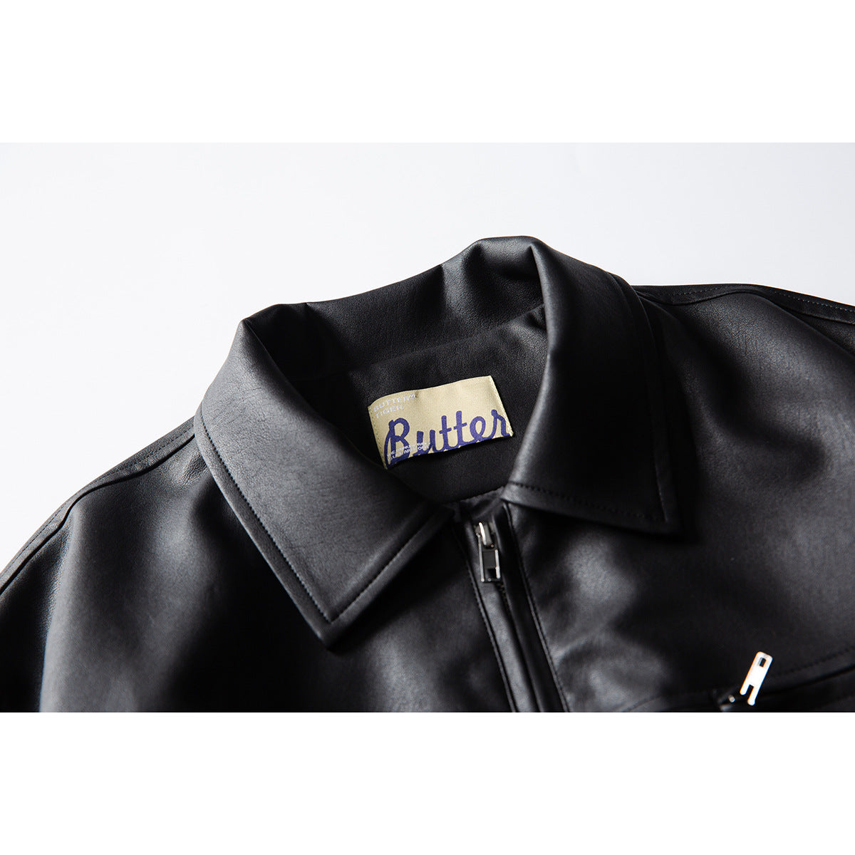 Close-up of a black jacket's pointed collar, center zipper, and cream Butter neck label.