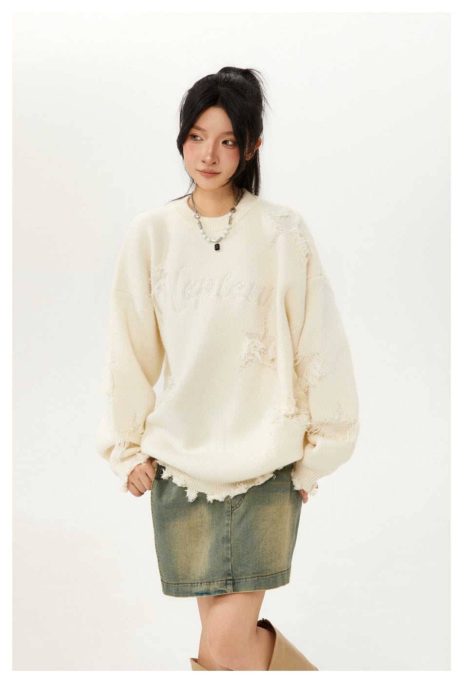 Closer front view of a model in an ivory sweater with distressed patches and tonal lettering, paired with a faded denim