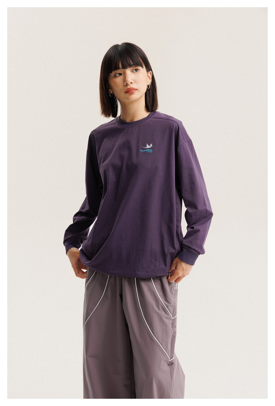 Model wearing a dark purple long-sleeve shirt with a small chest emblem and loose gray pants with white piping.