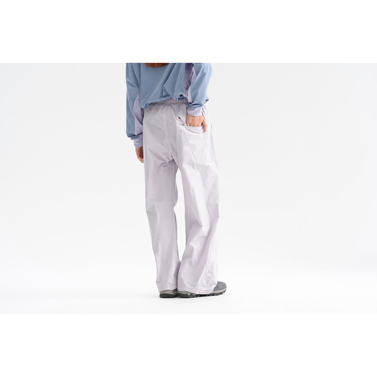 Rear view of a model wearing pale lavender loose trousers with a back pocket and gray shoes.