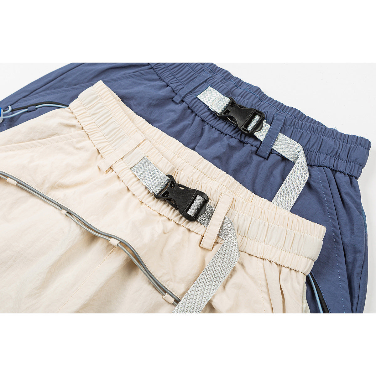 Close-up of beige and slate-blue pants showing elastic waistbands, patterned webbing belts, black buckles and drawcords.