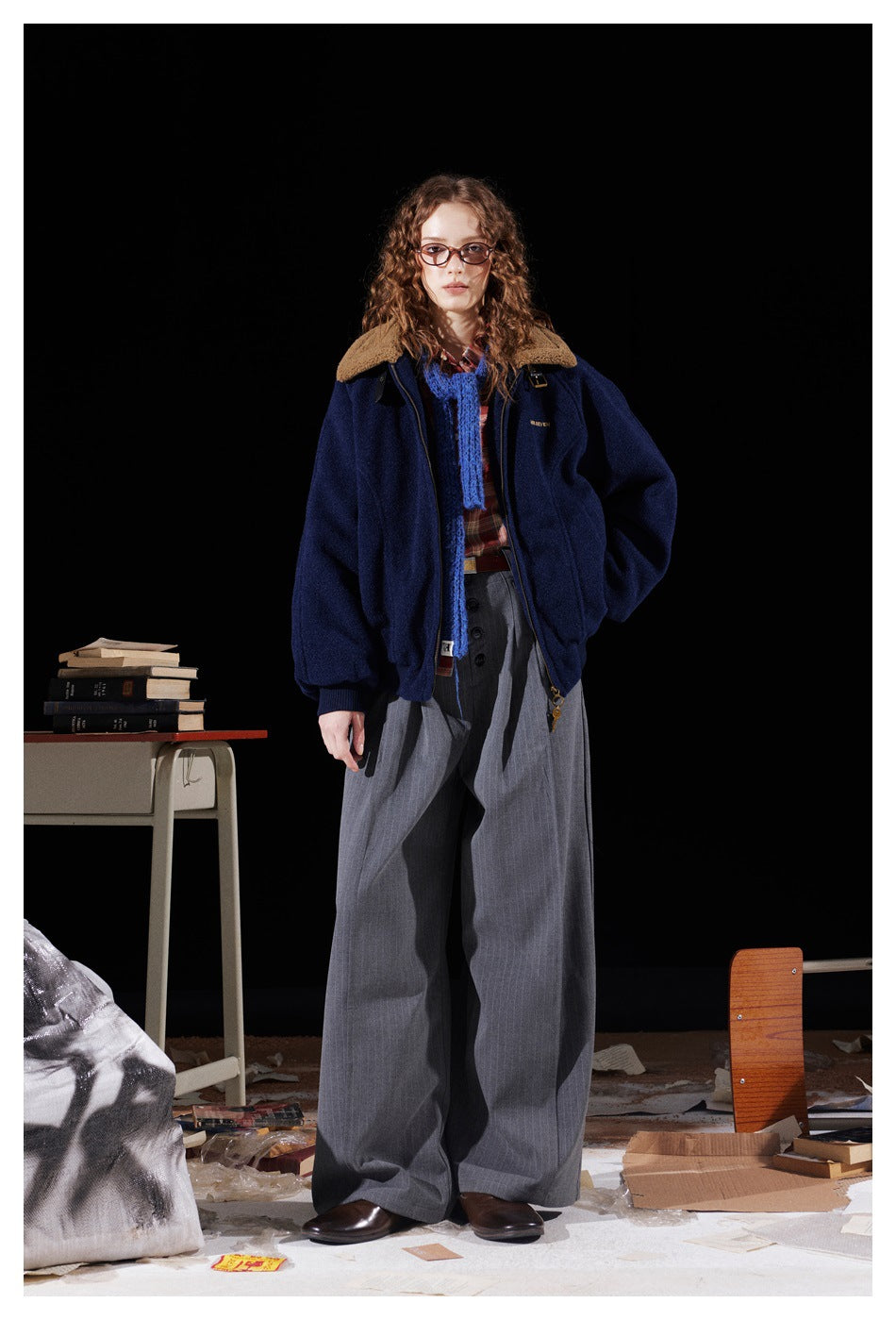 Full-length model wearing a navy jacket with a tan sherpa collar and wide gray trousers against a black backdrop.