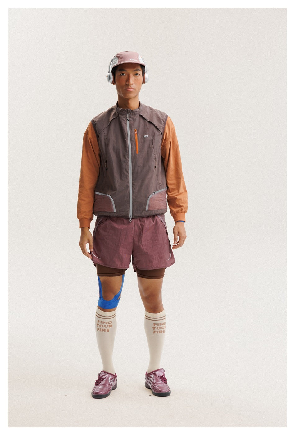 Full-body front view of a man wearing a zip-front taupe vest with orange sleeves and mauve shorts.