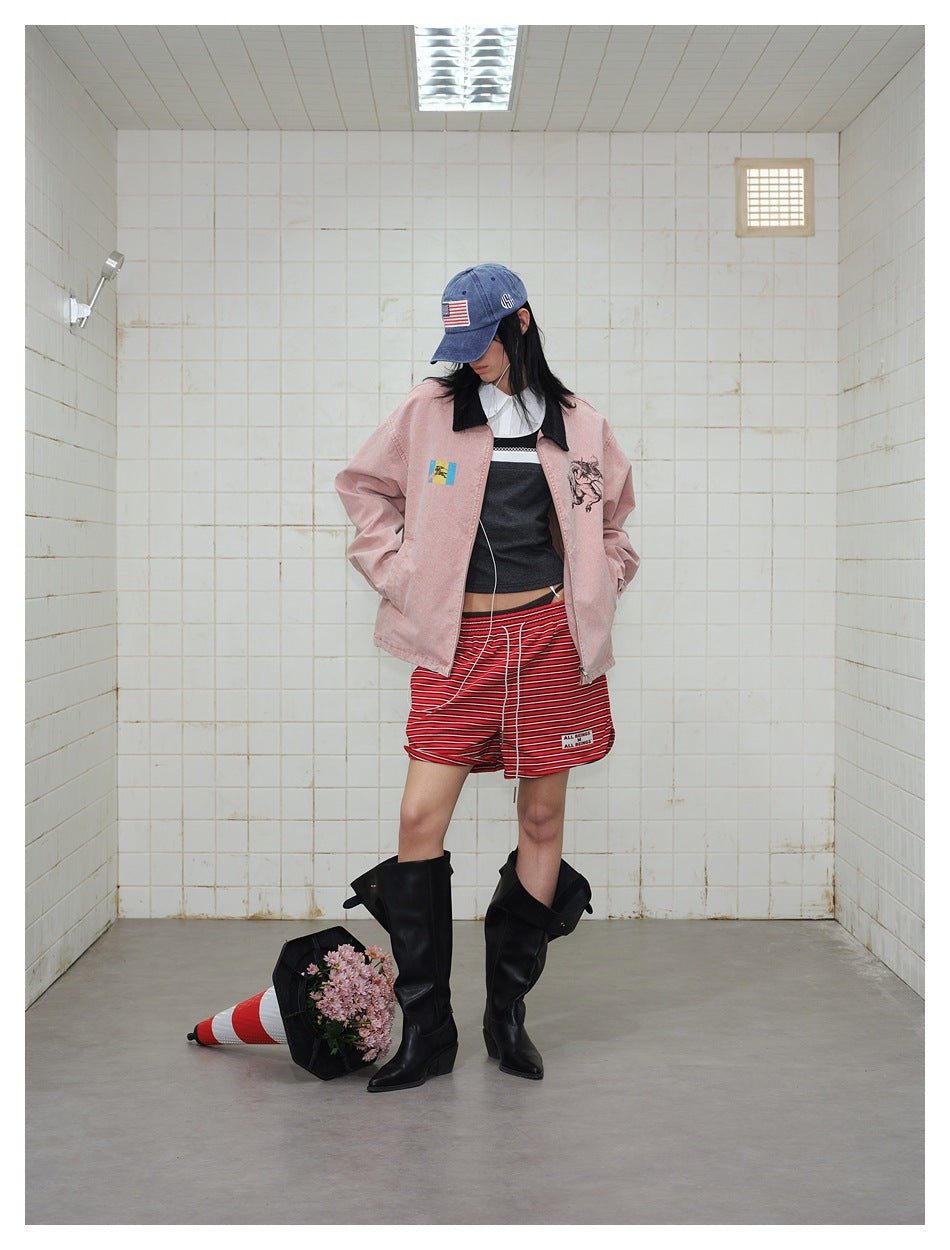 Model wearing an open dusty pink graphic jacket, red striped shorts, blue cap, and black boots, looking down.