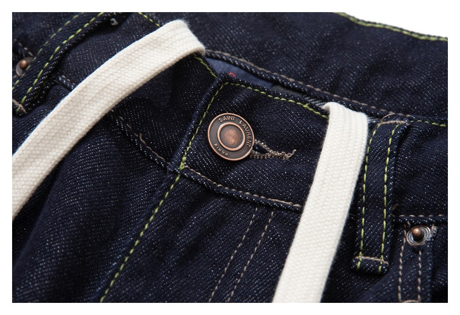 Jeans waistband close-up showing a branded metal button, belt loop, contrast stitching and white drawstrings.