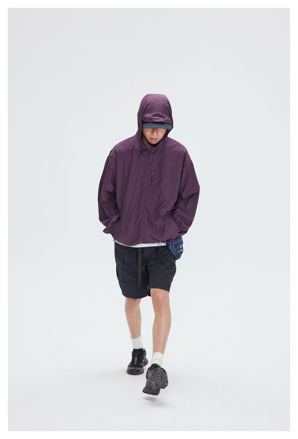 Model wearing a plum hooded jacket with a zip front and chest pocket, black shorts, and a cap.