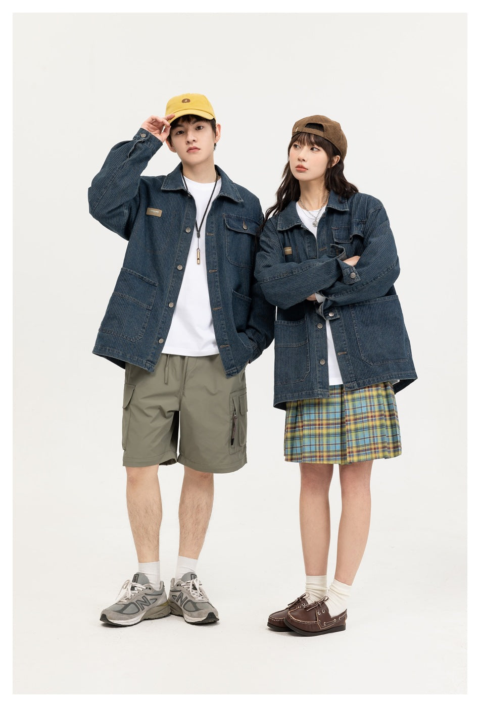 Two models pose in dark blue denim chore jackets with button fronts and multiple pockets, styled with shorts and a plaid