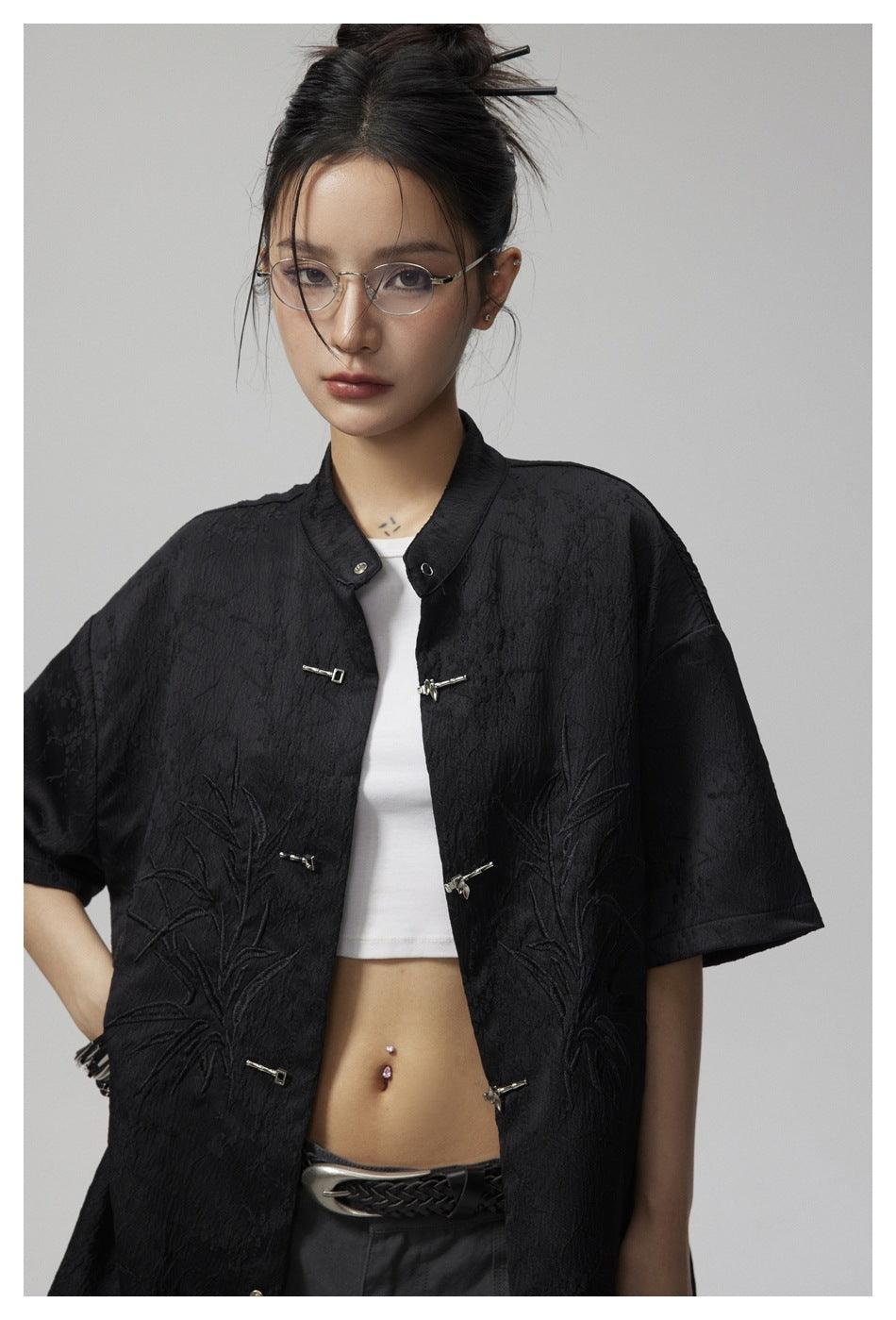 Close view of a model wearing a black jacket with short sleeves, metal frog closures and raised botanical embroidery.