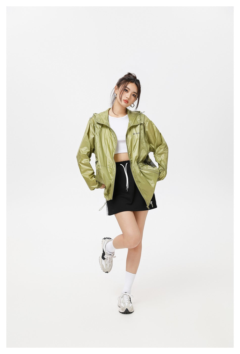 Model posing in an open olive-green hooded jacket with a white top and black skirt.