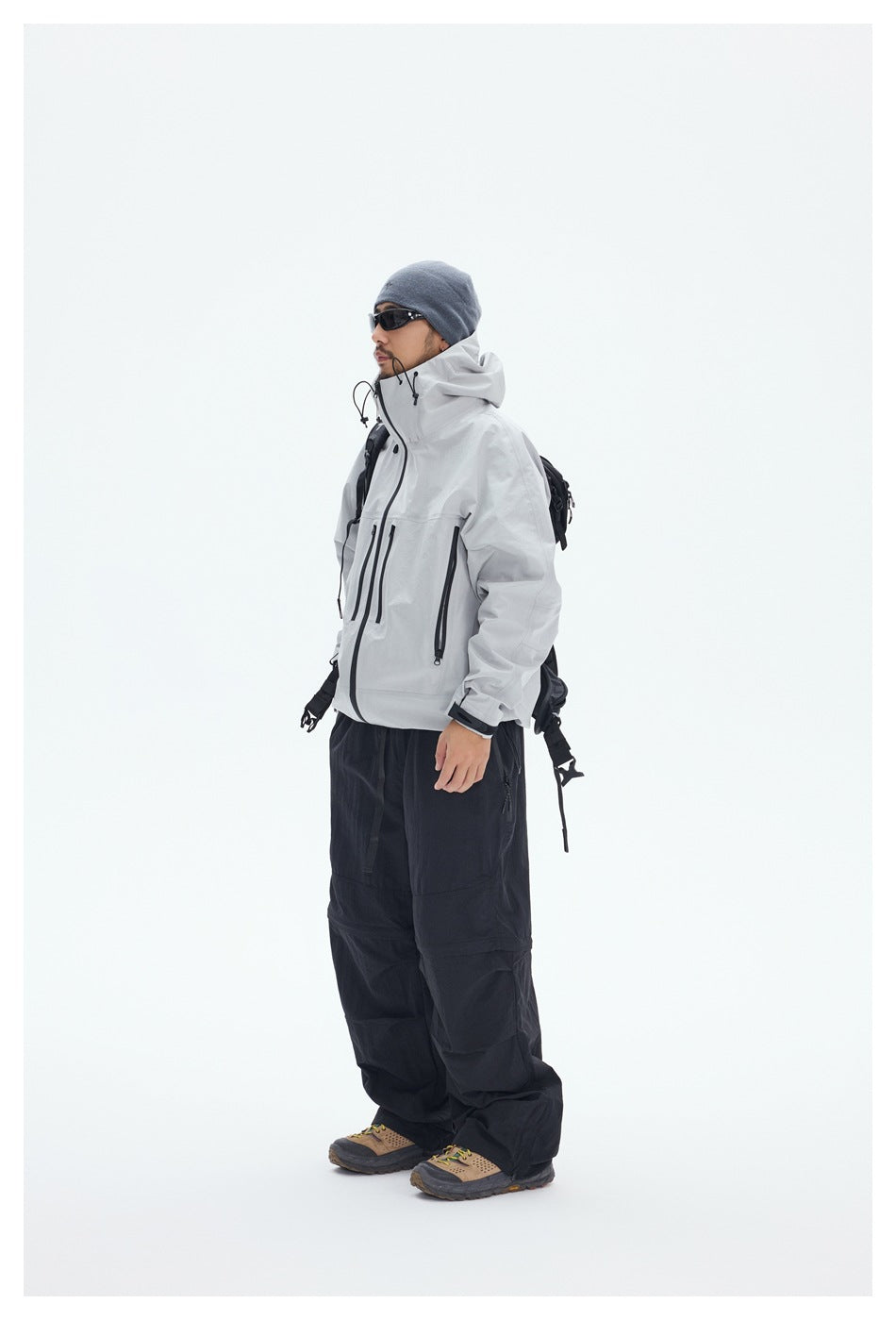 A man in a light gray hooded jacket and black pants is shown in side view wearing a hiking backpack.