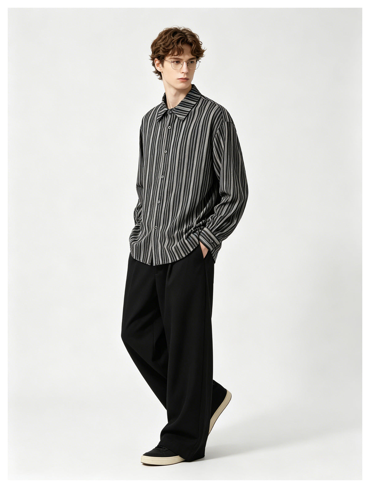 A man wears a dark button-up shirt with narrow gray vertical stripes, black trousers and sneakers.