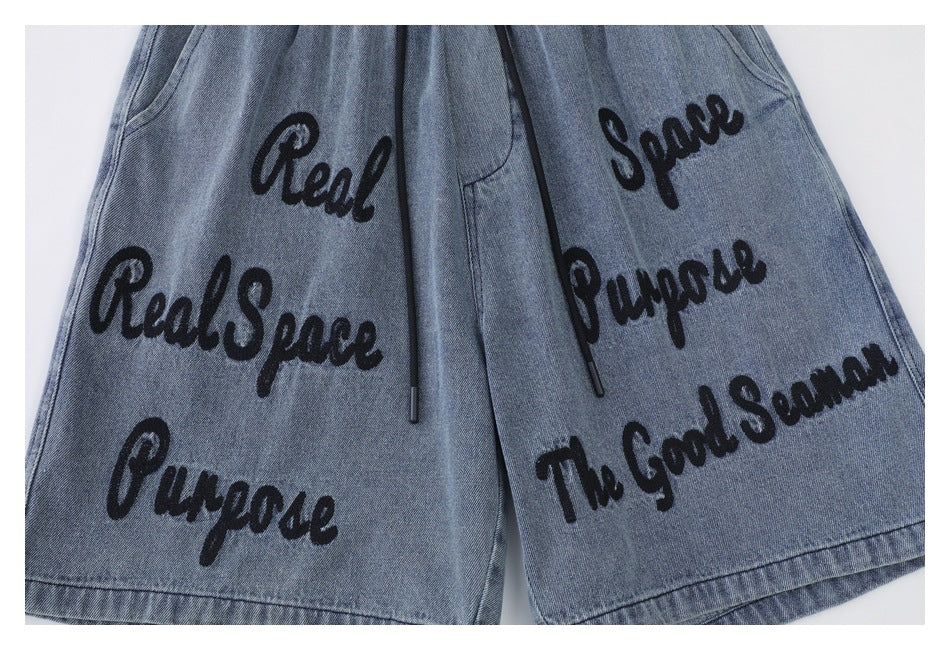 Front view of blue denim shorts with black drawcords and large black script graphics on both legs.