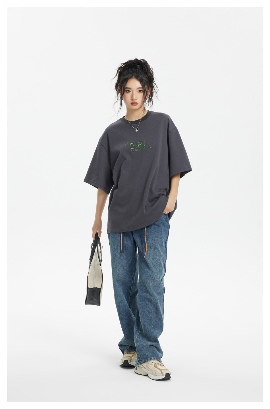 Model wearing an oversized charcoal T-shirt with green chest lettering and loose blue jeans.