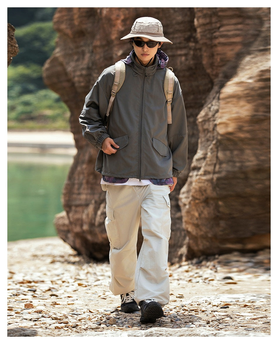 Person walking on a rocky shore in the dark gray hooded jacket with beige pants, backpack and bucket hat