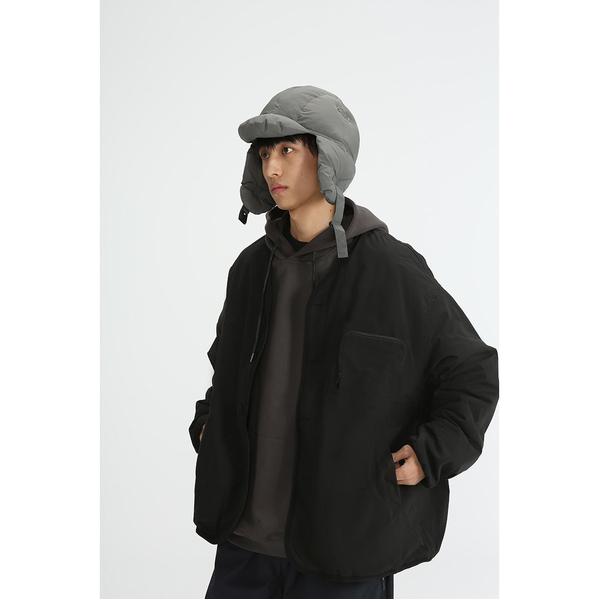 Three-quarter view of a model in a black oversized jacket with a chest pocket and a gray padded earflap cap.