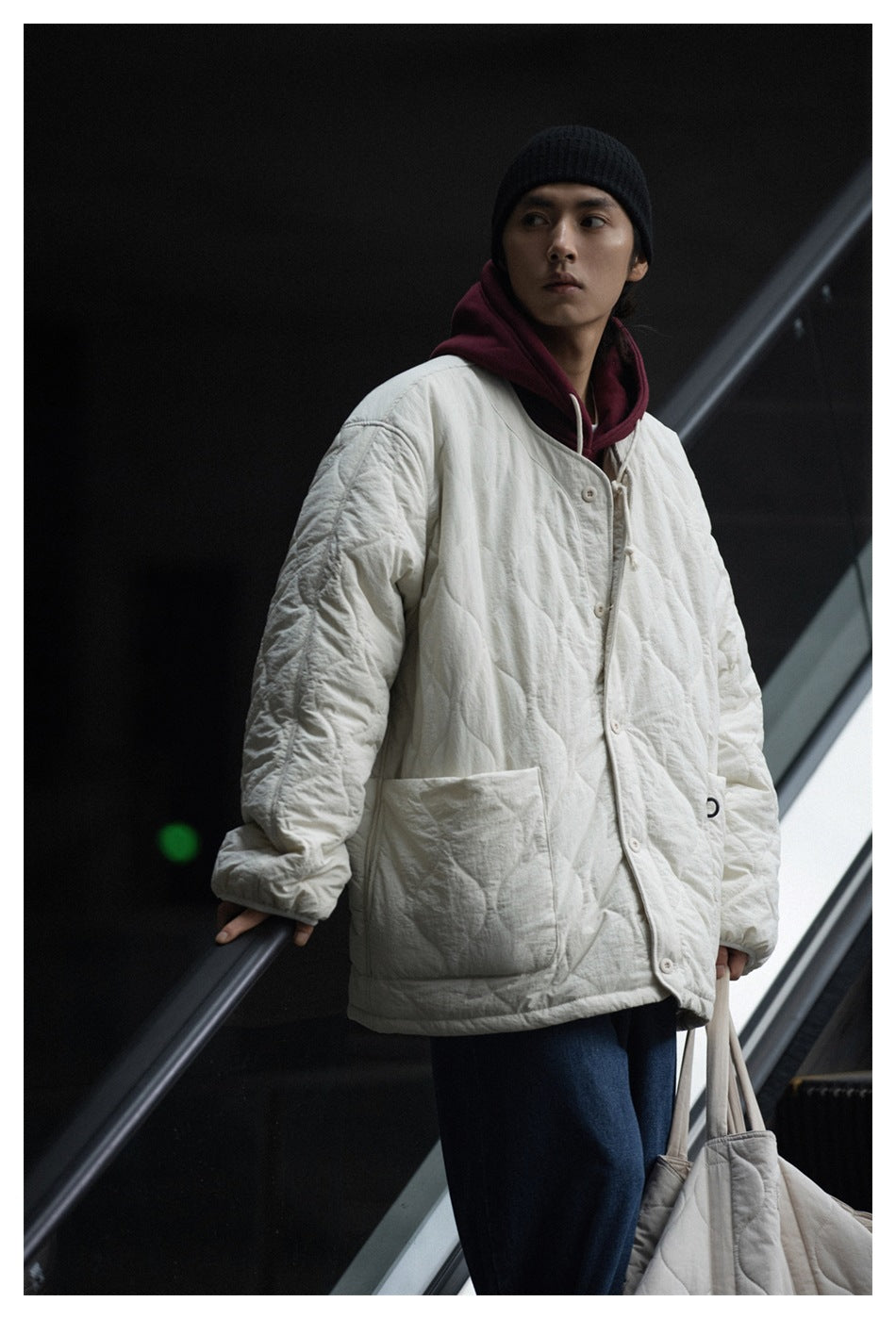 Person on an escalator wearing an off-white quilted jacket over a maroon hoodie with a black beanie and jeans