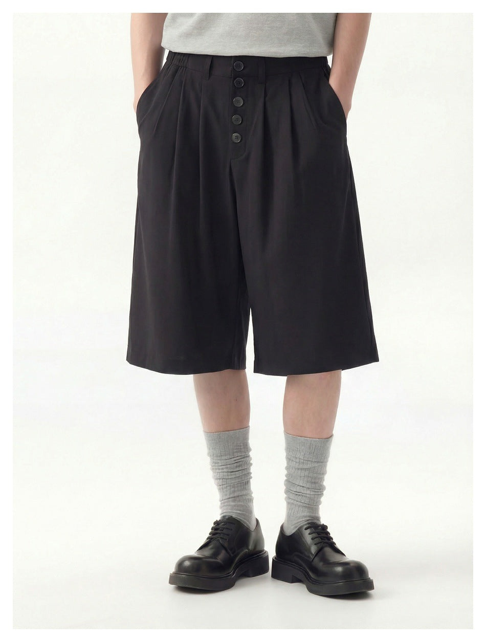 Front view of a model wearing black pleated knee-length shorts with a five-button fly and side pockets.