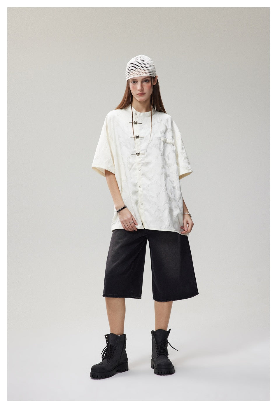 Model wearing a white patterned short-sleeve shirt with decorative frog closures, black knee-length shorts and a crochet cap.