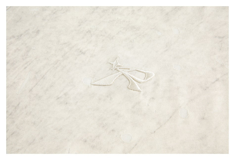Close-up of ivory mottled fabric with scattered distressed marks and a small embroidered star-like emblem.