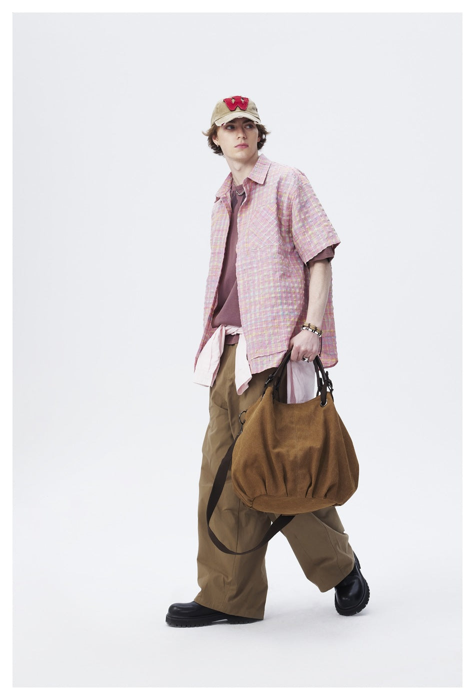 Model walking in a pastel pink plaid short-sleeve shirt and loose olive trousers, carrying a brown bag.