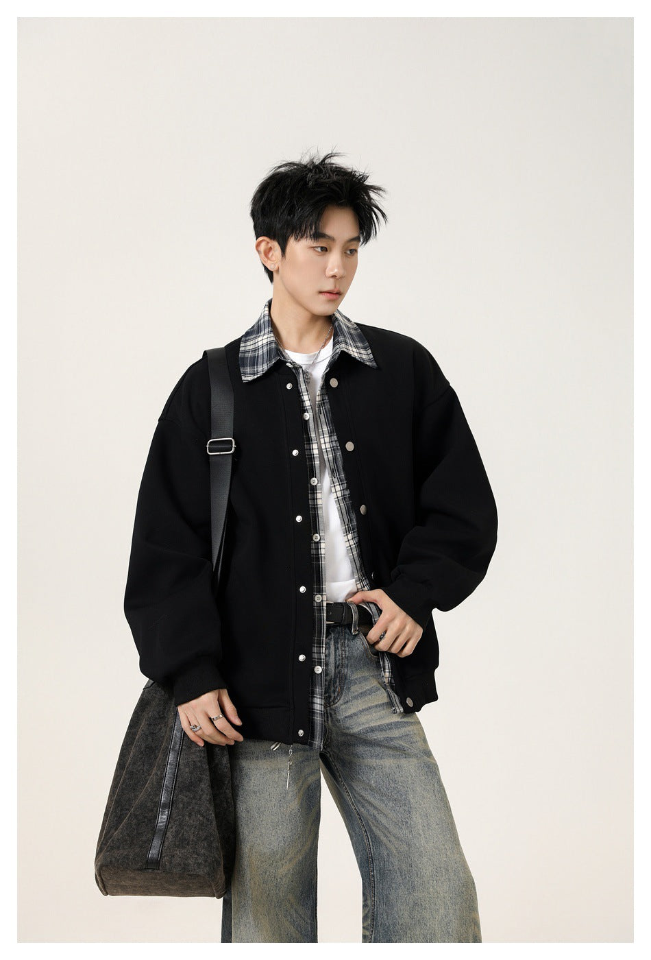 Model wearing an oversized black snap-front jacket with a gray plaid shirt, carrying a dark shoulder bag.