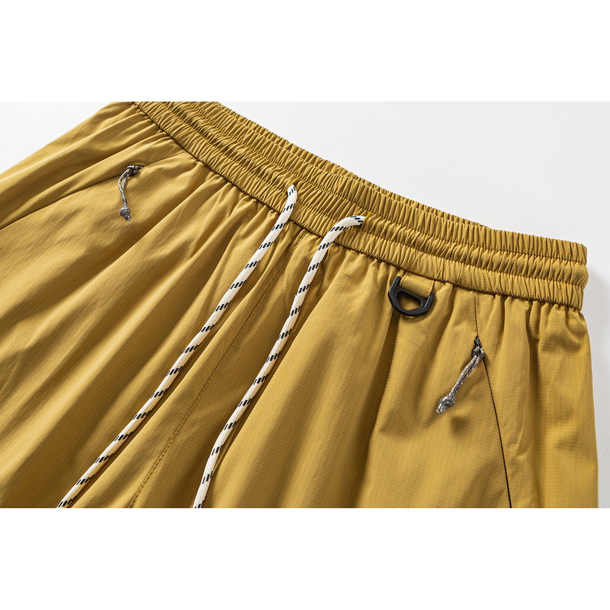 Close-up of mustard-yellow trousers showing the elastic waistband, white drawcords, and utility loop.