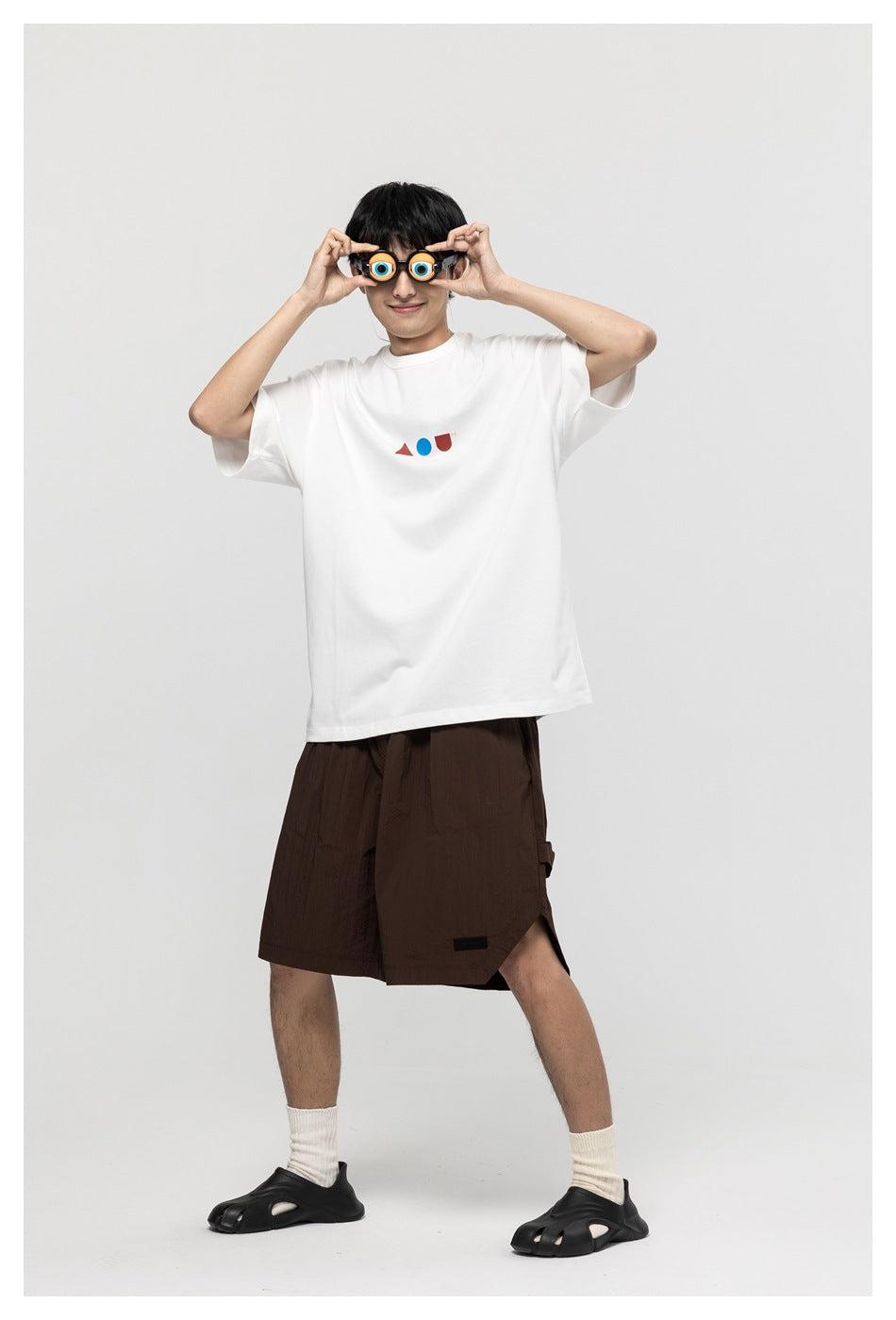 Model in white oversized T-shirt with small geometric chest print, brown shorts, white socks and black sandals, holding