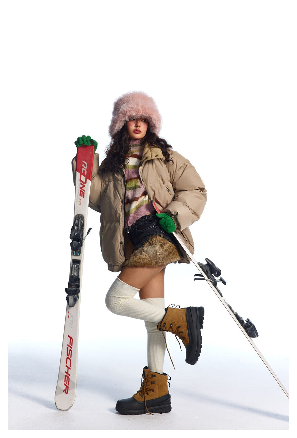 Model wearing a beige oversized puffer jacket, striped sweater and winter boots while posing with skis.