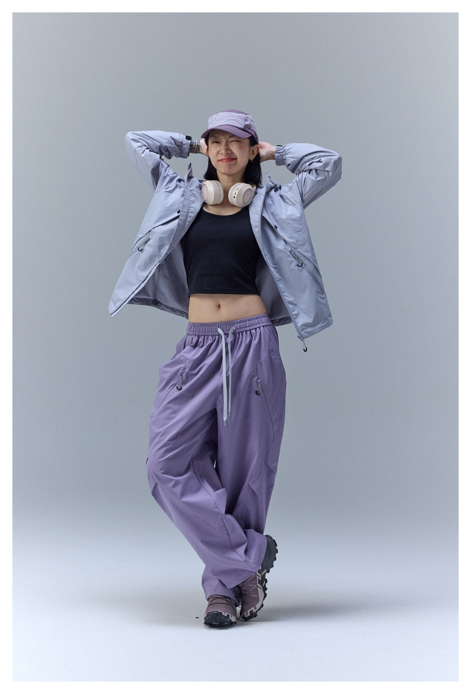 Model poses with a light gray jacket open over a black top and loose lavender drawstring pants.