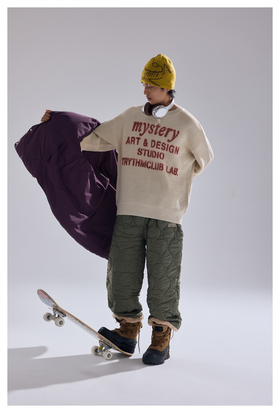 Model in a cream graphic sweater, olive quilted trousers, and yellow beanie holds a plum vest beside a skateboard.