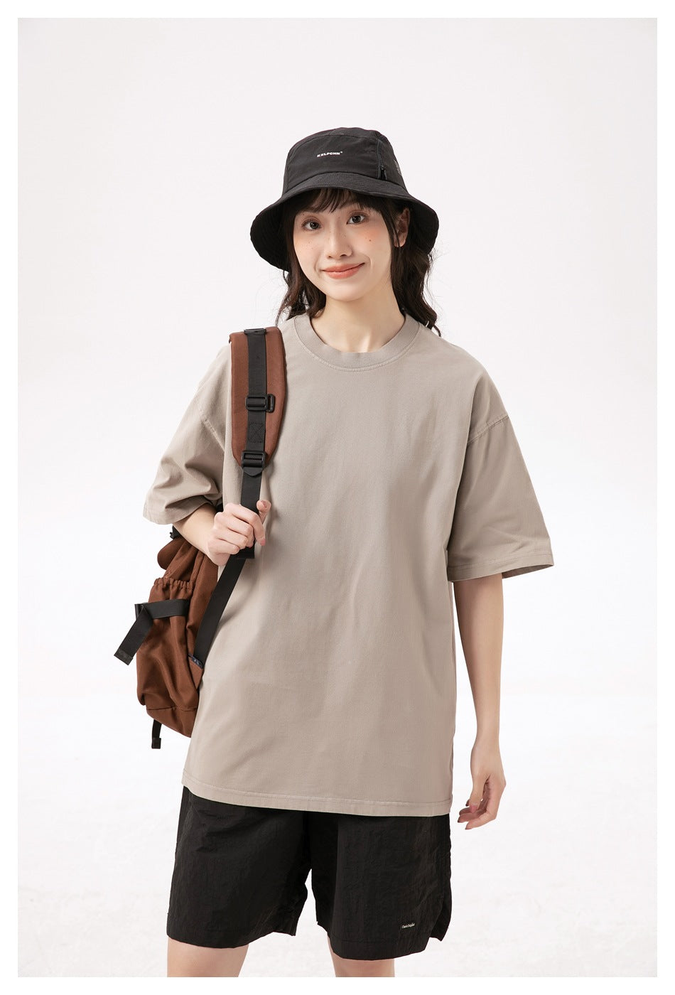 Model wearing a loose beige T-shirt and black shorts with a black bucket hat and brown backpack.