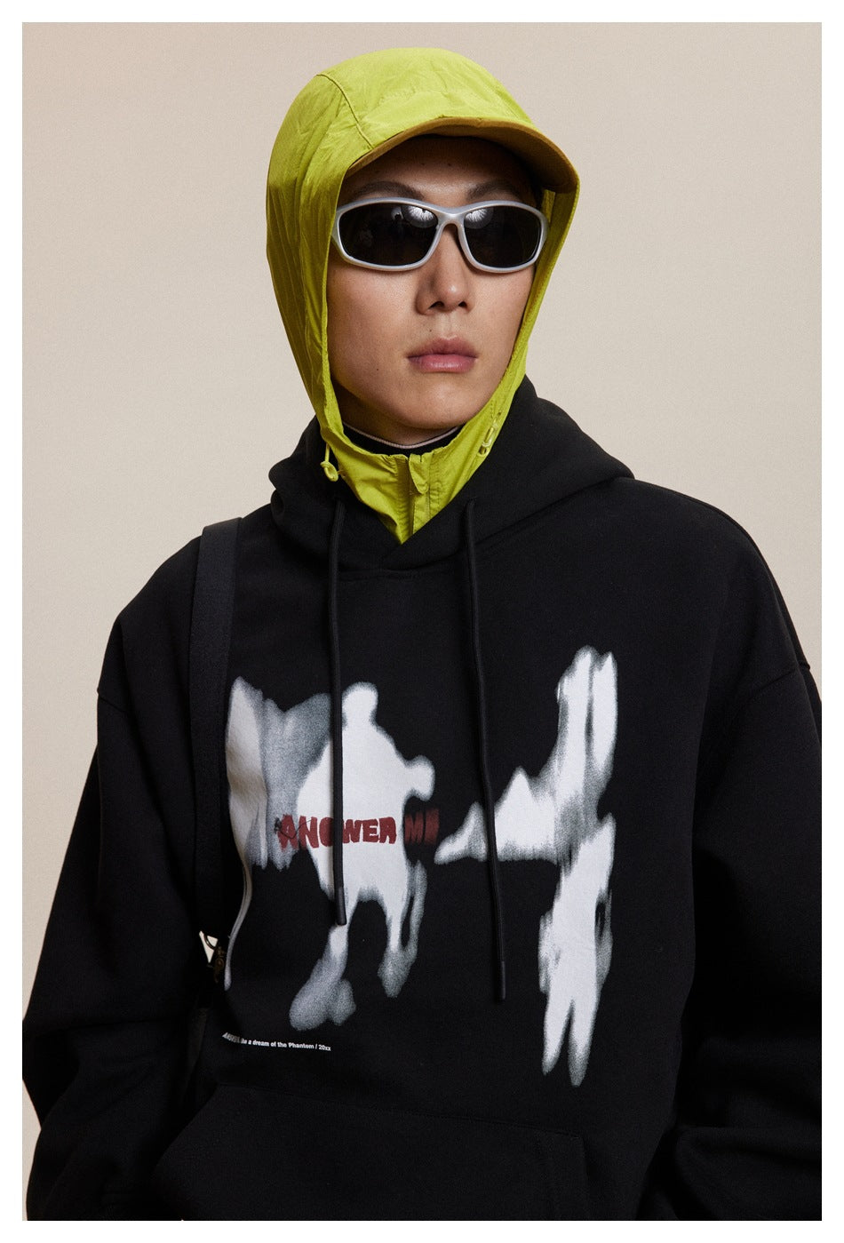 Front close-up of a black hoodie with a large white abstract graphic and red lettering, layered over neon yellow.