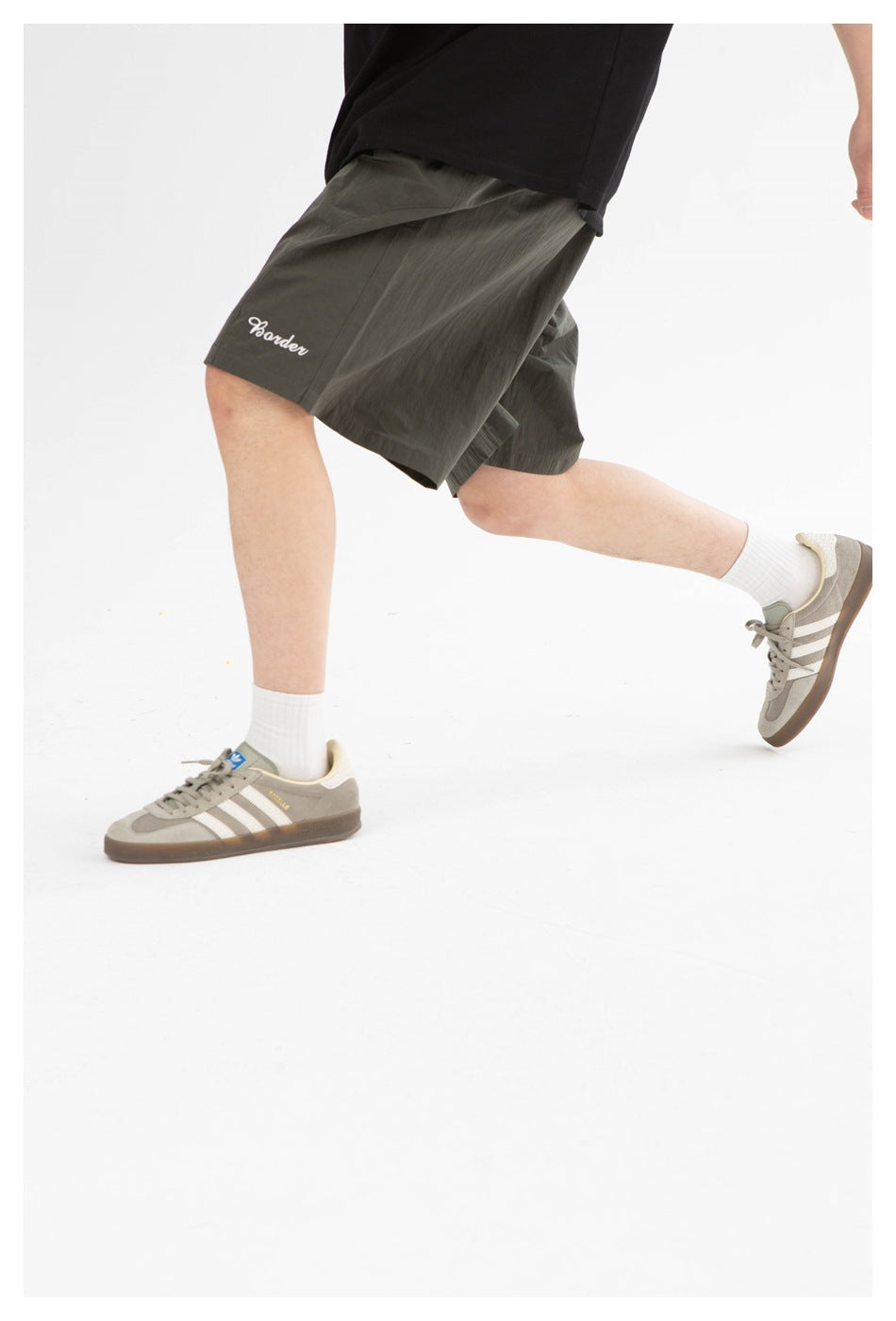 Person in olive shorts, black tee and gray sneakers with white socks, mid-stride, cropped from the waist down