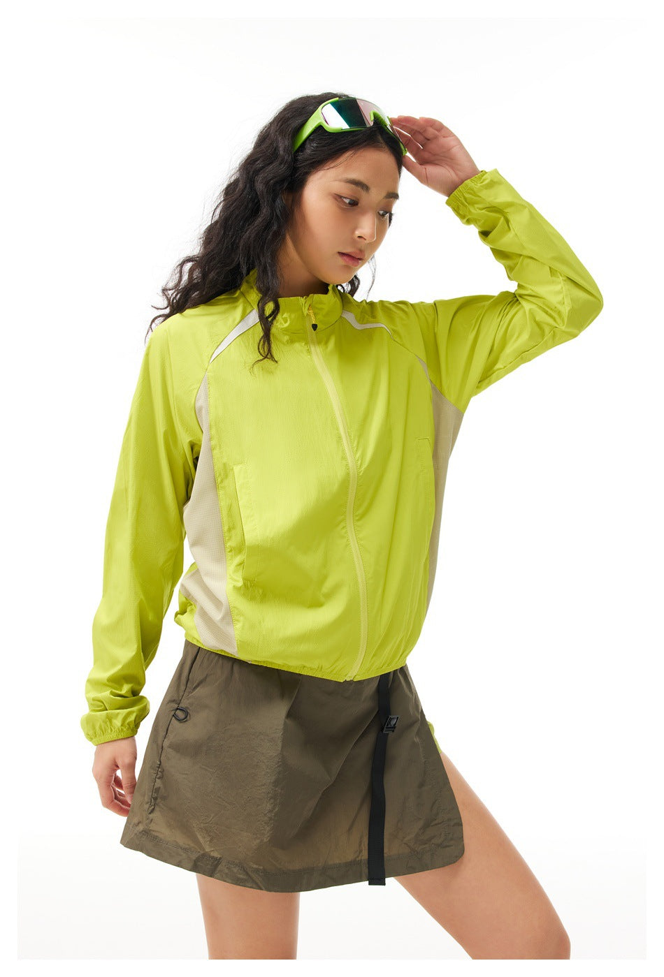 Woman wearing a yellow-green zip jacket with beige side panels, hand raised to her sunglasses