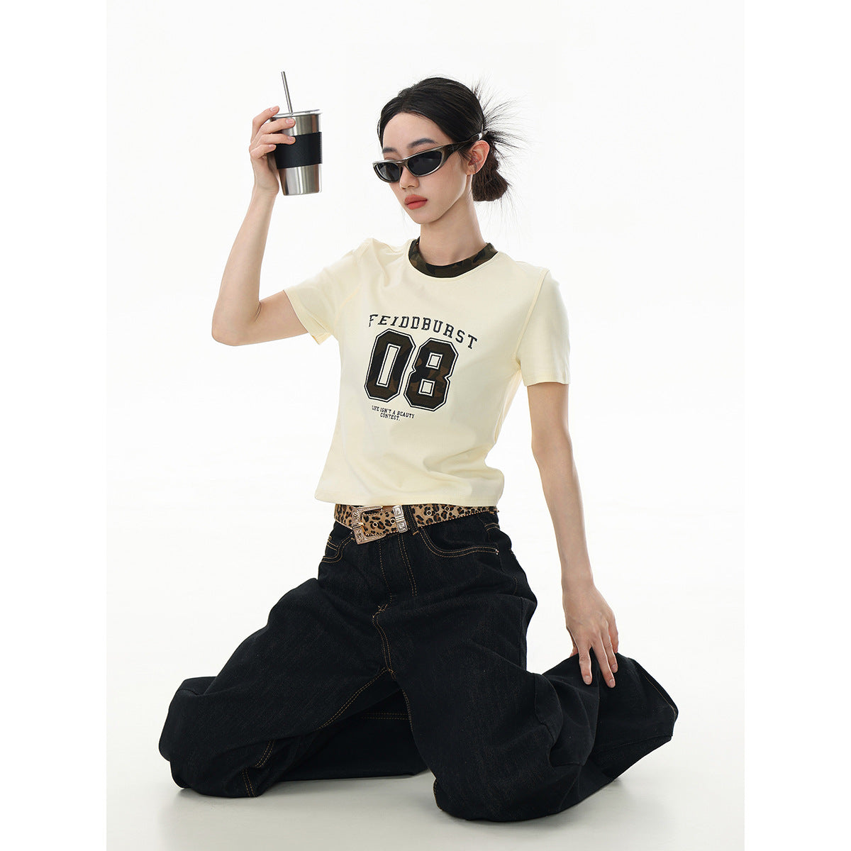 Model kneeling in an ivory graphic T-shirt and dark wide-leg jeans, holding a tumbler; front view.