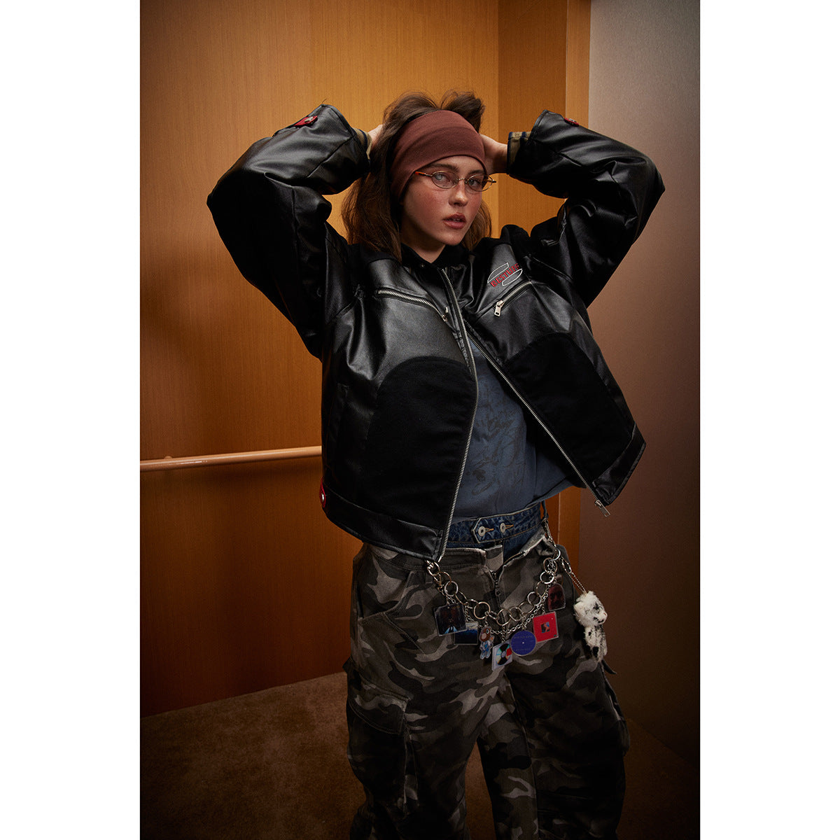 Model wearing a black glossy zip-front jacket with chest zippers and camouflage trousers.