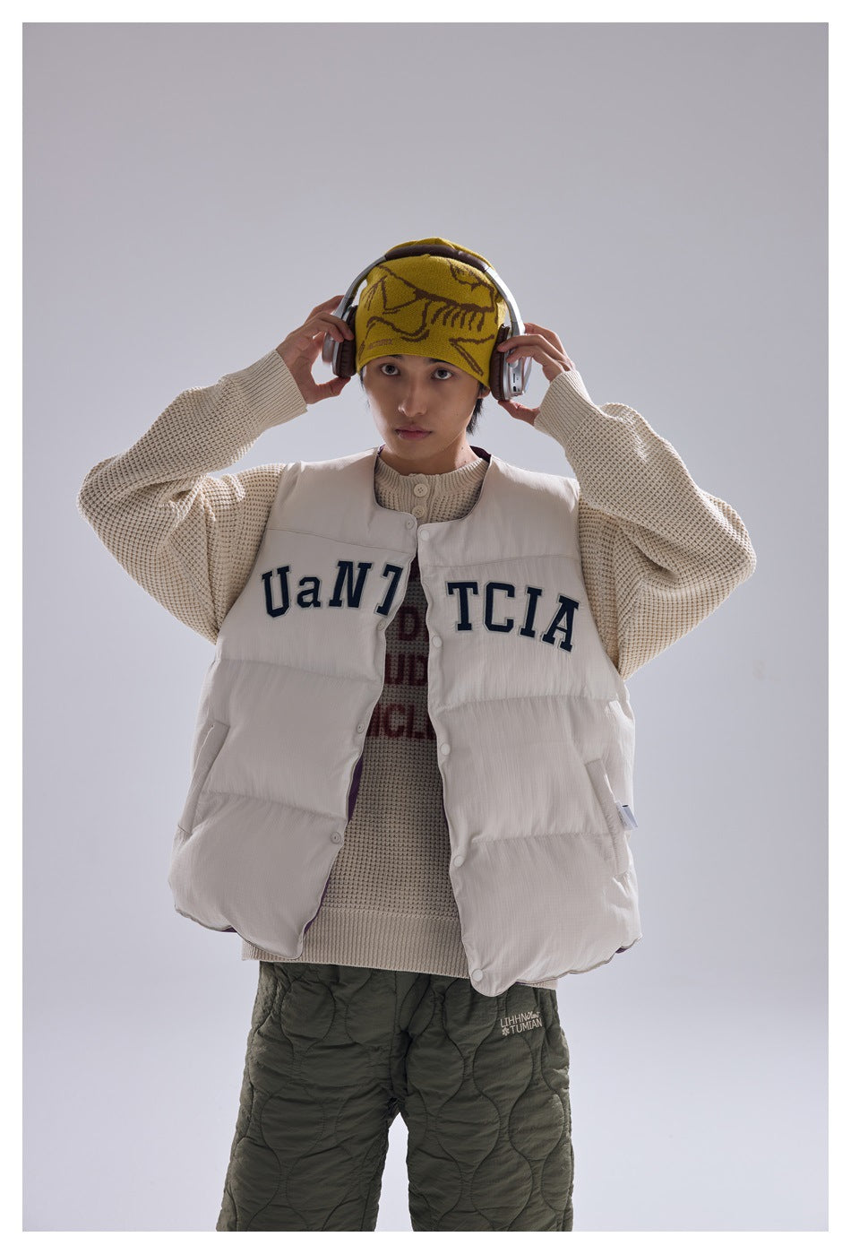 Person wearing off-white puffer vest over a beige waffle knit sweater with olive quilted pants, hands on headphones