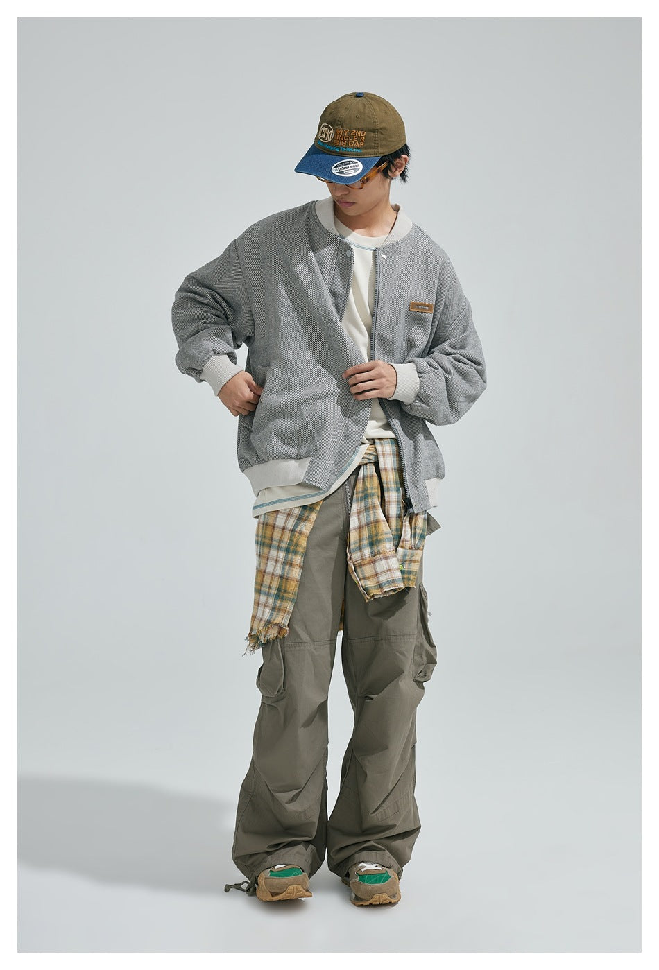 Full-length model wearing a gray herringbone bomber jacket with olive cargo pants and a plaid shirt tied at the waist.